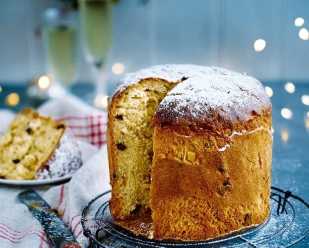 Image of panettone set on a festive table with fairy lights. The bread has a slice cut out of it