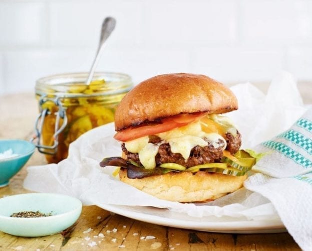 Pickles shown in a glass jar behind a large burger in a bun