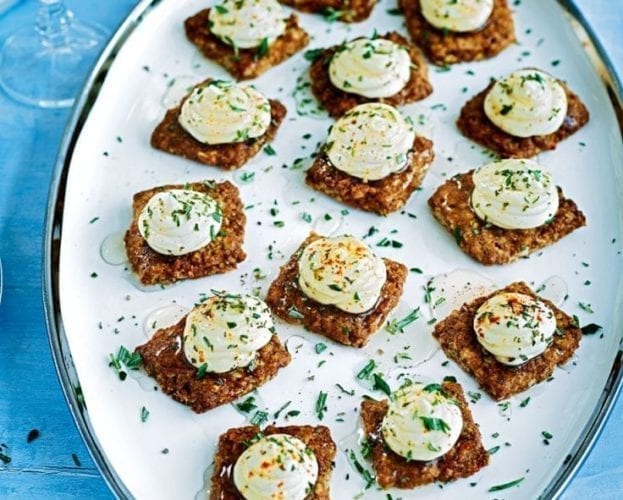 Lots of oat biscuits topped with goat's cheese and herbs shown on an oval plate