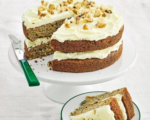 Iced carrot cake shown on a white cake stand, with a slice cut out