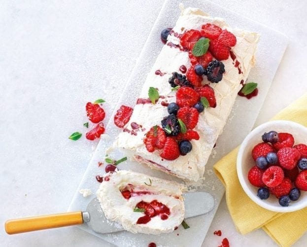 A meringue roulade shown on a board, with a slice taken out. It's topped with berries
