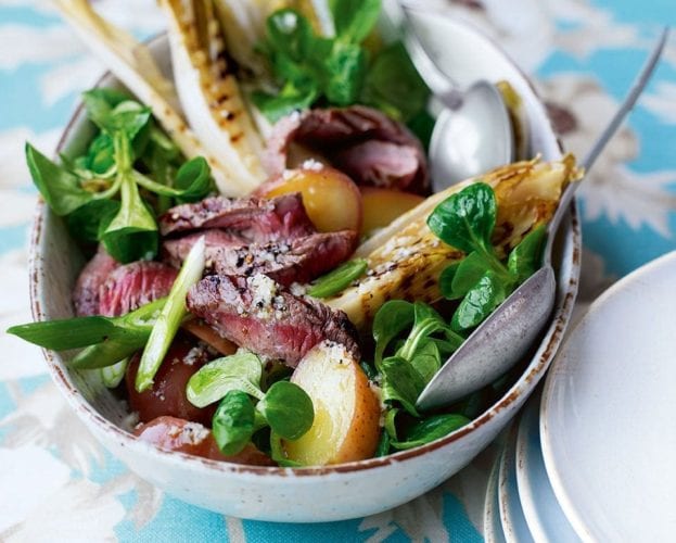 A colourful steak and potato salad shown in a bowl on a floral tablecloth