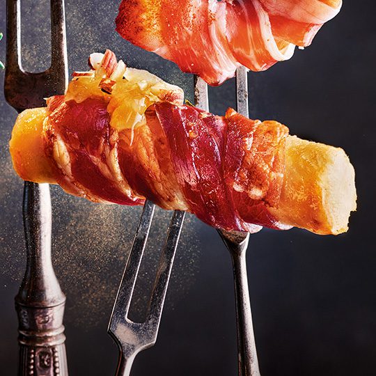 A baton of celeriac wrapped in bacon speared on a fork