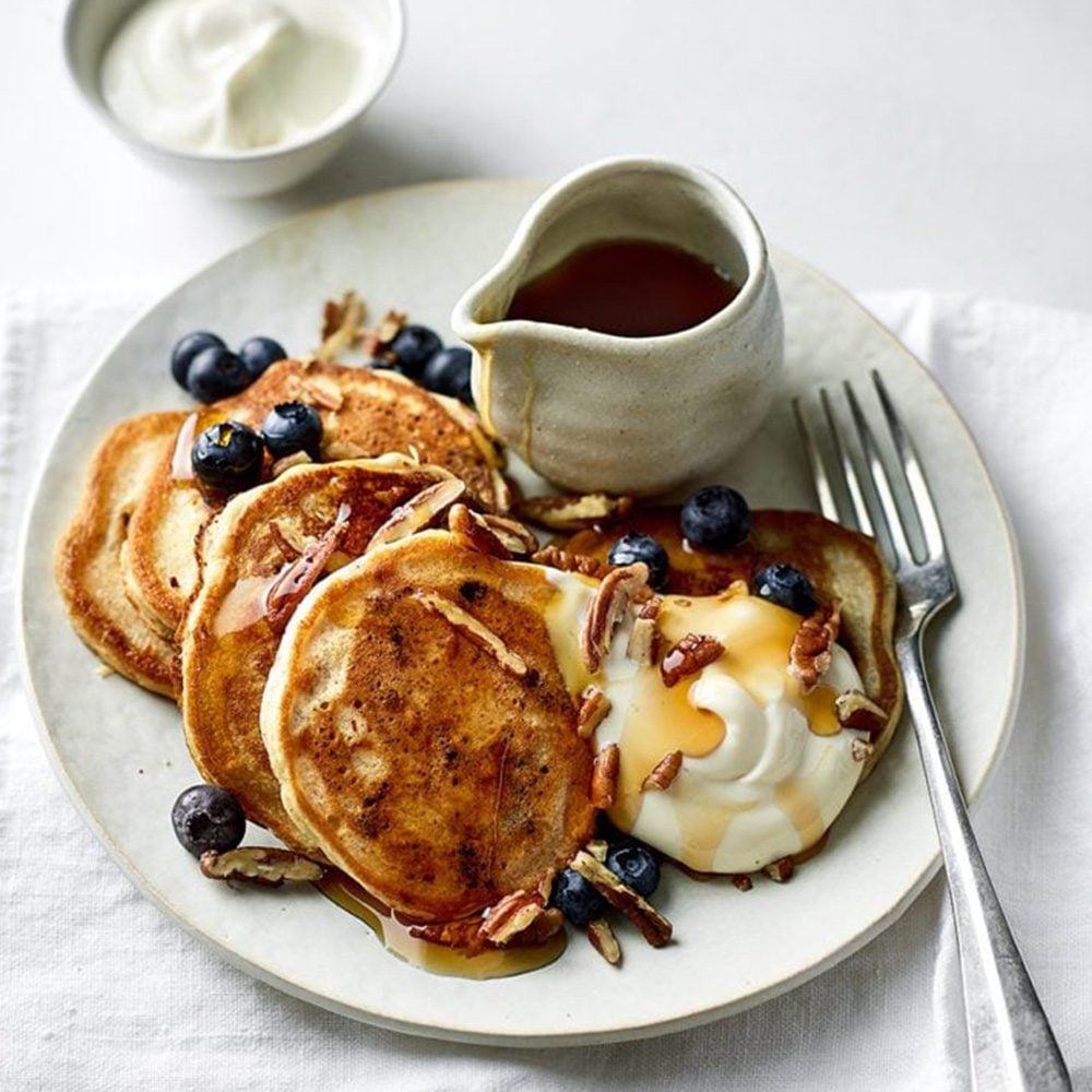 A pile of vegan pancakes topped with blueberries, maple syrup and vegan yogurt, with more maple syrup on the side