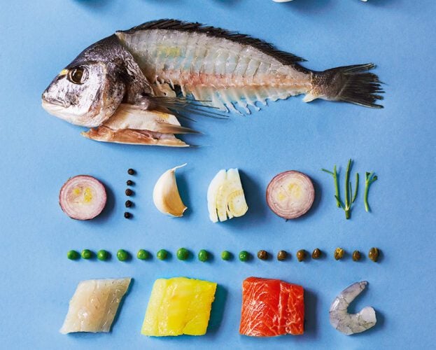 The ingredients of a fish pie laid out together, including a whole filleted fish, spinach, potatoes, prawns and soft herbs