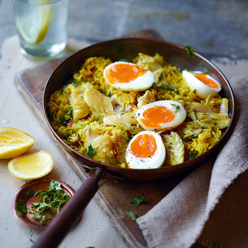 A pan of kedgeree with halved soft boiled eggs and lemon wedges on the side