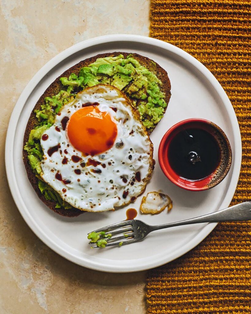 A plate with toast topped with avocado and a fried egg, and a small bowl of soy sauce on the side
