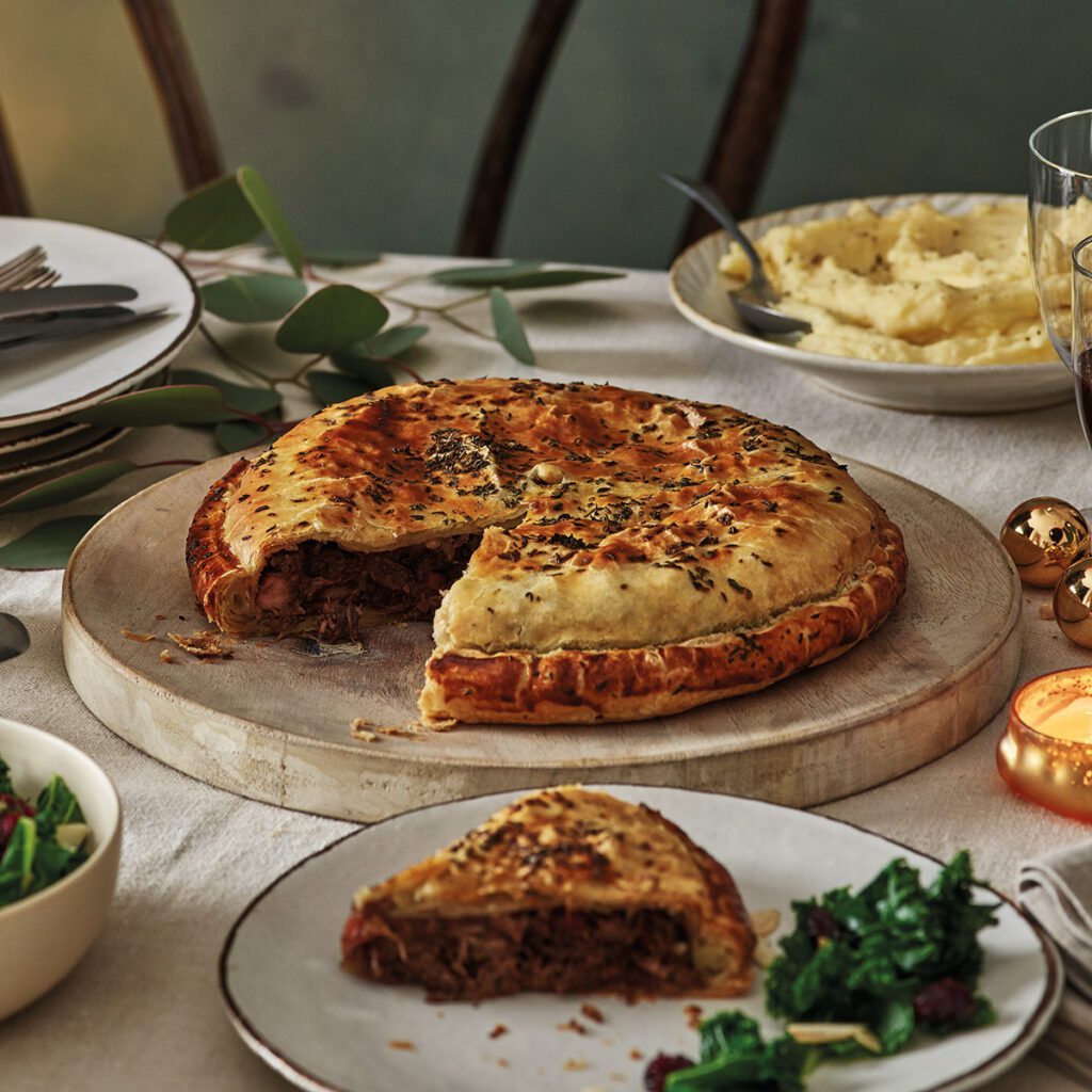 A rustic pastry pithivier on a board, with a slice on a plate in the foreground