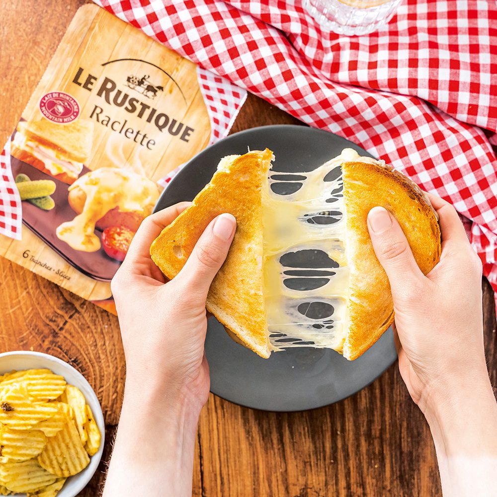 Two halves of a cheese toastie being pulled apart, with strings of cheese in between, and a packet of raclette cheese slices in the background