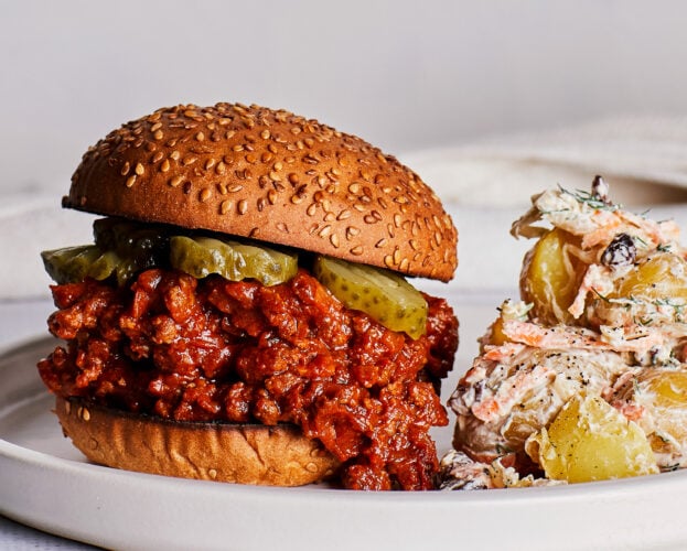 A sloppy joe bun topped with pickles, with coleslaw potato salad on the side
