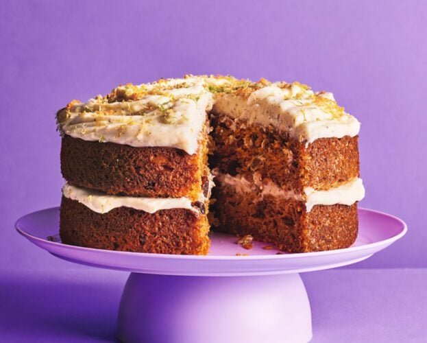 A large carrot cake, topped with citrus zest, on a purple cake stand