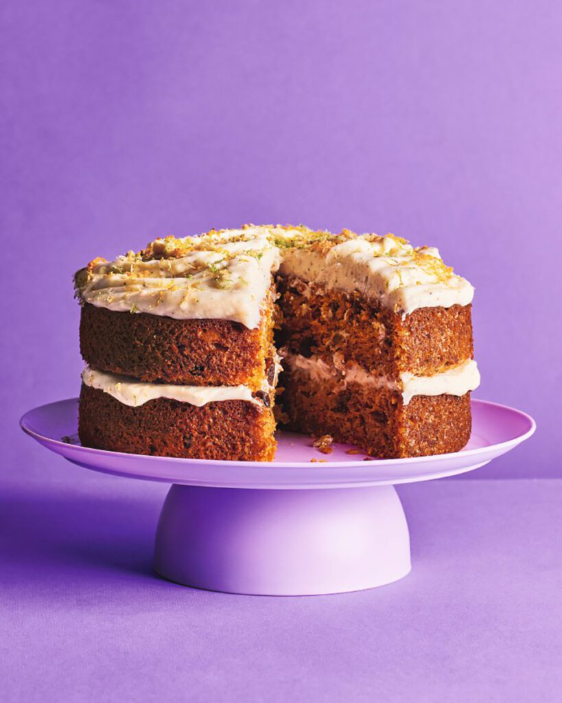 A large carrot cake, topped with citrus zest, on a purple cake stand