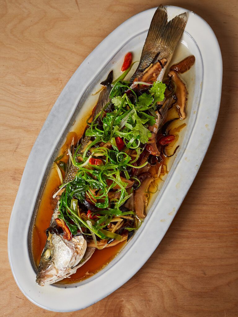 A patter with a whole steamed sea bass, topped with spring onions and herbs