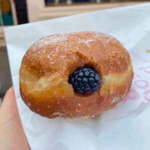 A doughnut dusted in sugar and with a blackberry lodged where is has been filled with bramble jam