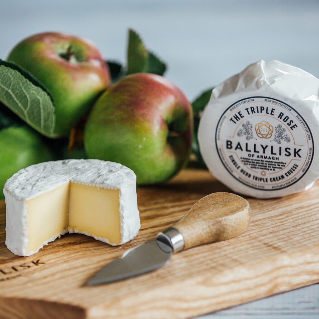 Ballylisk Triple Rose cheese cut open on a board, with a packaged cheese and apples in the background