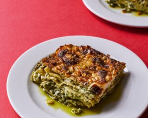 A slice of wild garlic pesto lasagne on a white plate in the foreground, with another plate of lasagne in the background