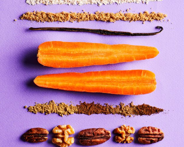 The ingredients to make a carrot cake, including carrots, a vanilla pod, flour, sugar, sultanas and nuts, laid on a purple background